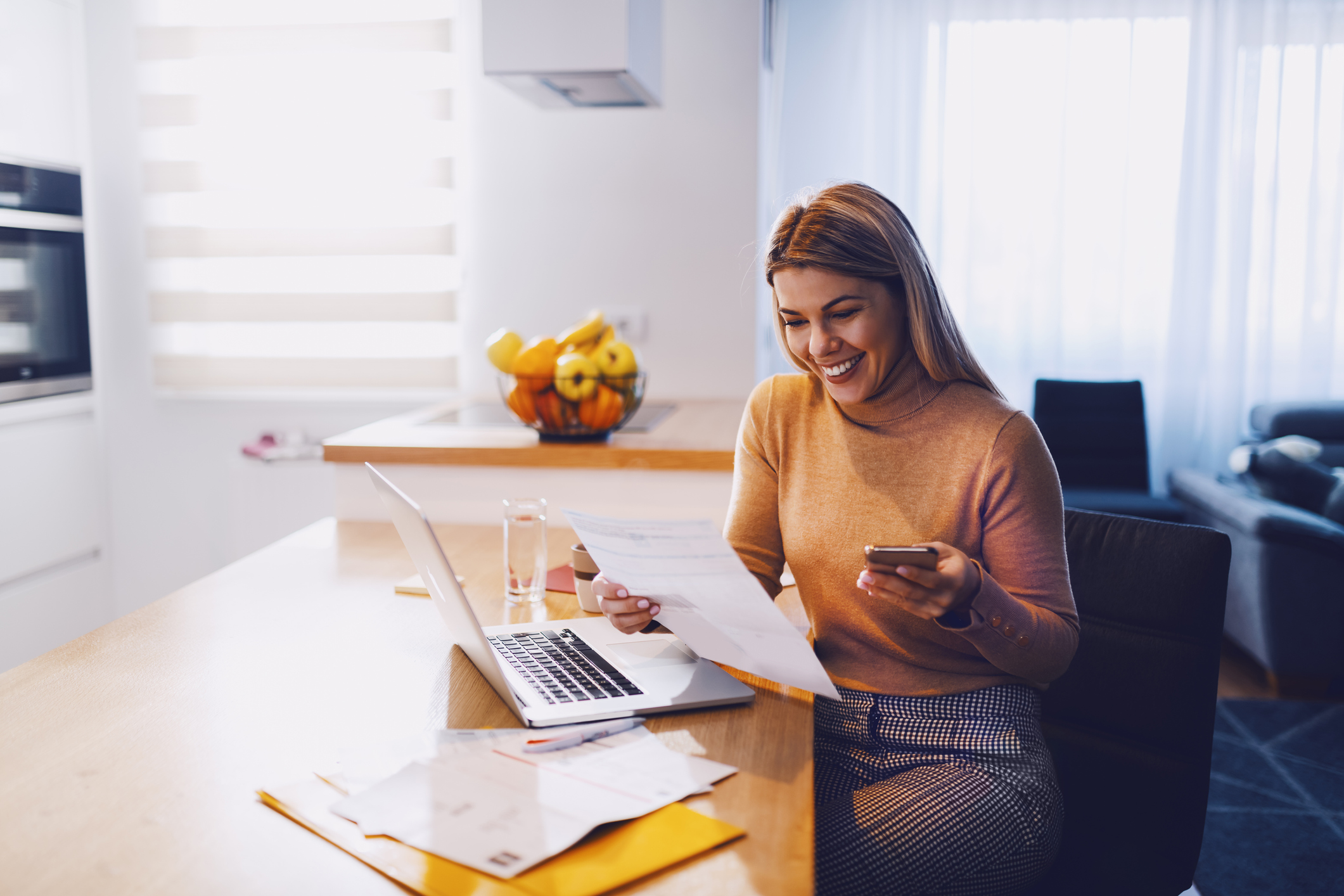 woman in sweater holding bills in one hand and in other smart phone. On table are laptop and bills